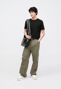 T-shirt preta de mangas curtas usada com calças cargo verde-oliva com múltiplos bolsos, combinada com sapatilhas brancas e bege e uma mala preta.