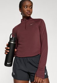 Maroone Halbzip Langarmtop mit grauem Logo, kombiniert mit schwarzen elastischen Shorts mit grauem Logo. Die Person hält eine schwarze Wasserflasche.
