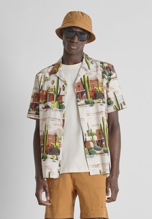 Man met een beige vissershoed, zwarte zonnebril, wit t-shirt, kort mouw blouse met woestijnthema en mosterdkleurige korte broek, staand tegen een lichte achtergrond.