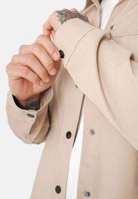 Camisa beige de botones con una tela ligera, botones a presión negros y detalle en los puños. Tatuajes visibles en la mano.