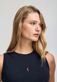 Granatowy top bez rękawów z okrągłym dekoltem, z małym czerwonym haftowanym logo z przodu. Materiał wydaje się miękki i gładki.