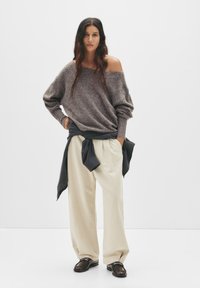 Oversized brown knit sweater, cream wide-leg trousers, and black shoes. A dark jacket tied around the waist adds contrast.
