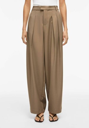 Kvinde iført brune bukser med høj talje og brede ben med plisseringer, kombineret med sorte thong-sandaler og en hvid ærmeløs top.