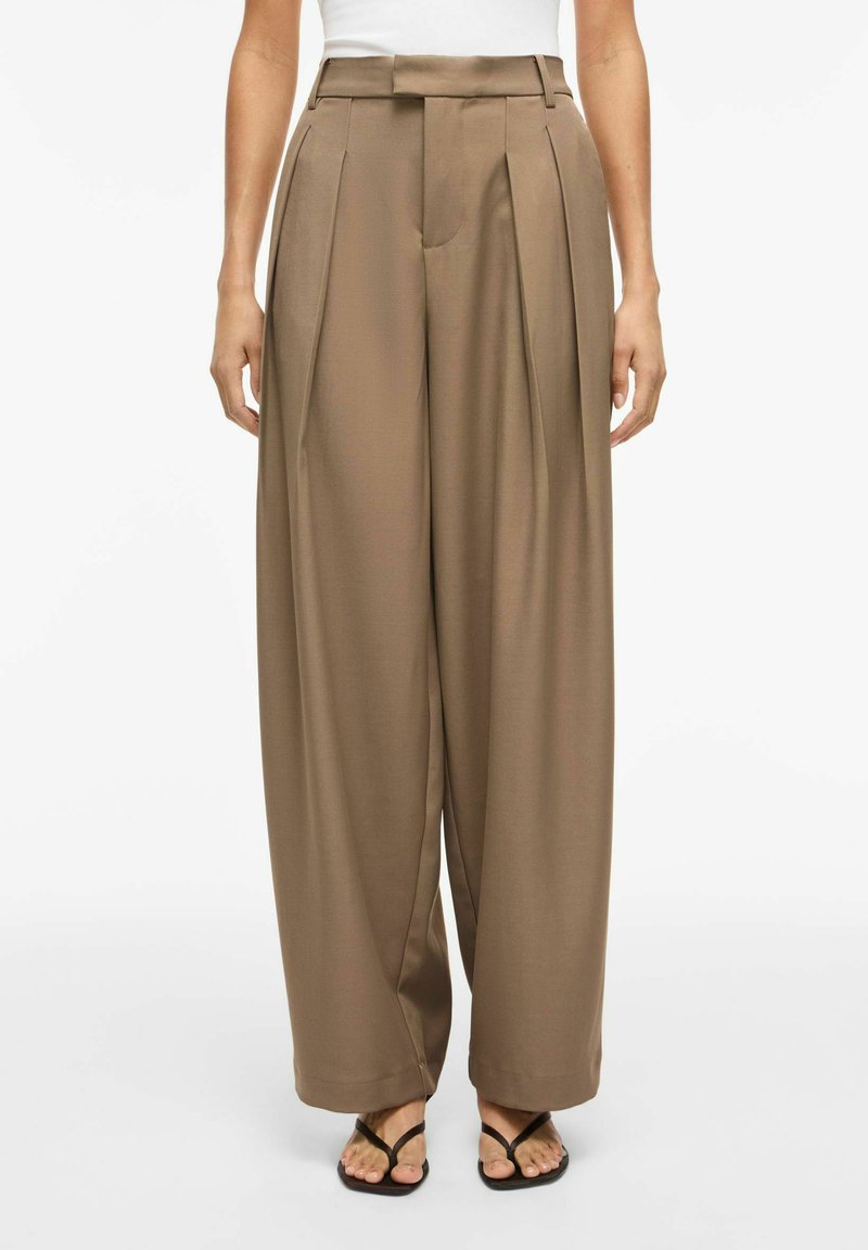 Γυναίκα που φοράει καφέ παντελόνες με υψηλή μέση και φαρδιά πόδια με πτυχώσεις, συνδυασμένες με μαύρα σανδάλια τύπου σαγιονάρα και λευκό αμάνικο τοπ.