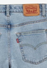 Los jeans de mezclilla azul claro cuentan con una etiqueta de cuero marrón, una etiqueta roja y costuras amarillas en contraste. Incluyen dos bolsillos traseros con costuras decorativas.