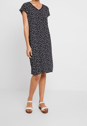 Kobieta w czarnej sukience do kolan z nieregularnym, białym wzorem w kropki oraz białych sandałach na kostkę z odkrytymi palcami.