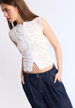 Jeune femme portant un haut en dentelle blanche sans manches avec des fermetures à boutons grenouille et un jean large taille haute bleu foncé, mains dans les poches, regardant à gauche.