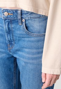 Lyseblå denimjeans med en synlig lomme og brun søm, sammen med en kremfarget genser med lange ermer som har en avslappet passform.