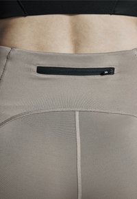 Beige Sporthosen mit glatter Textur, versehen mit einer schwarzen Reißverschlusstasche auf der Rückseite und subtilen Nähten entlang der Nähte.