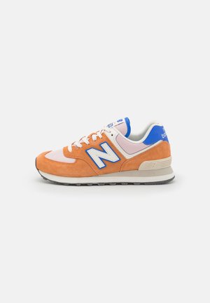 Pomarańczowo-różowy sneaker New Balance z białymi sznurówkami, niebieskim logo "N" oraz niebieskim detalem na pięcie na jasnoszarym tle.
