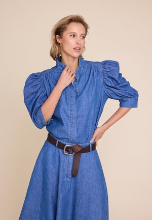 Kvinne iført blå denimkjole med puff-ermer, høy krage og brunt belte, stående med en hånd på hoften og ser til siden.