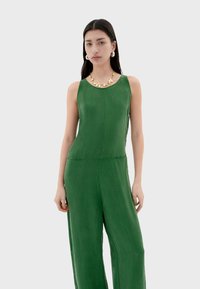 Combinaison verte sans manches, fabriquée dans un tissu lisse. Présente un col rond et des poches latérales. Les accessoires comprennent un collier jaune et blanc.