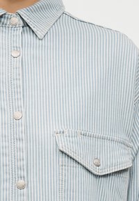 Πουκάμισο με κάθετες ρίγες σε ανοιχτό μπλε και λευκό, με ασημένια κουμπιά κλικ και τσεπάκι με καπάκι στο στήθος.