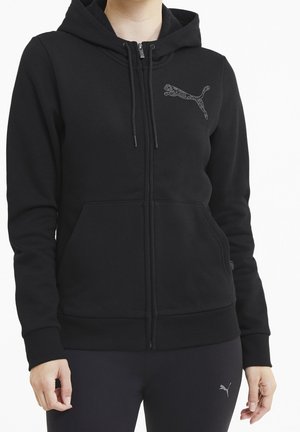Kobieta ubrana w czarną bluzę Puma z kapturem i zamkiem błyskawicznym oraz czarne legginsy z logo Puma na udzie.