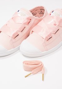 Paire de baskets en toile rose avec de larges rubans en satin en guise de lacets et un lacet rose supplémentaire enroulé devant sur une surface blanche.