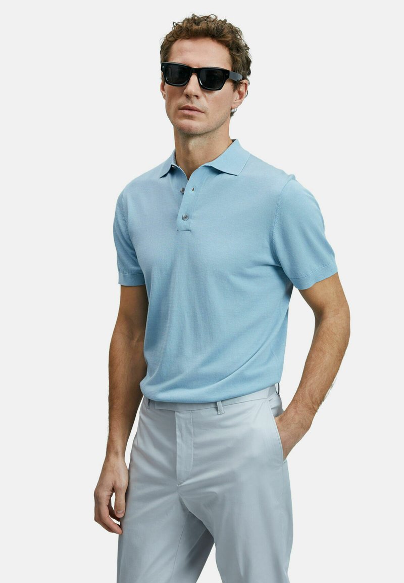 Helles blaues Polohemd mit kurzen Ärmeln, einem Kragen und drei Knöpfen, kombiniert mit hellgrauen Hosen. Das Modell trägt schwarze Sonnenbrillen.