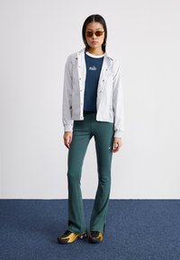 Lys grå jakke med trykknapper over en marineblå Nike t-skjorte, kombinert med mørkegrønne flared bukser og svarte/gylne sneakers.