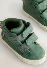 Zöld bőrből készült magas szárú cipők, két tépőzáras pánttal, texturált felülettel és fehér gumitalppal. Oldalukon mintázott díszítéssel.