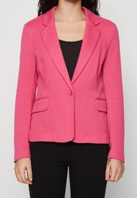 Rosa kavaj i texturerat tyg, med spetsig krage, enkel knappstängning och två framfickor. Bärs över en svart topp.