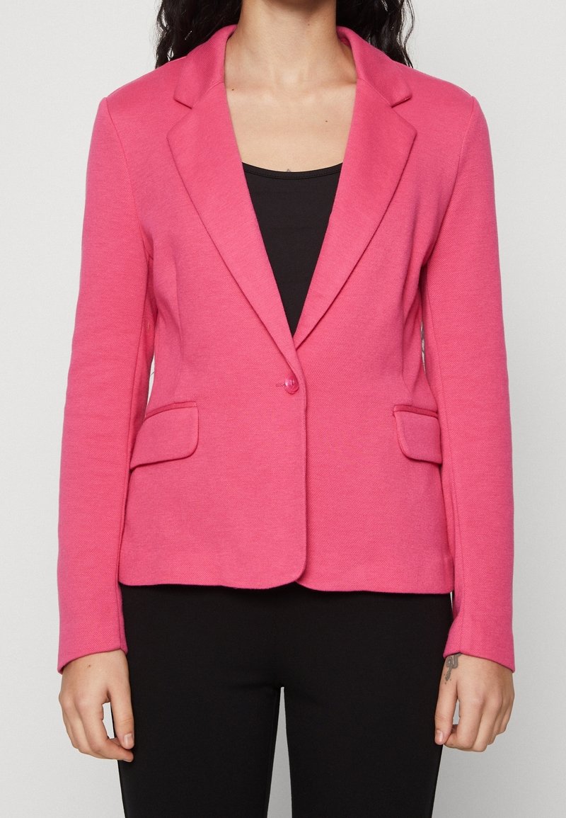 Rosa kavaj i texturerat tyg, med spetsig krage, enkel knappstängning och två framfickor. Bärs över en svart topp.