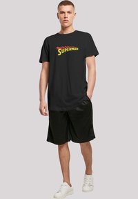 Black cotton t-shirt featuring a yellow "SUPER" text design with red accents. Paired with black shorts and white sneakers.