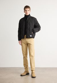 Čierna prešívaná bomber bunda so zipsom, v kombinácii so svetlohnedými chinos nohavicami a béžovými členkovými čižmami. Štruktúrovaná tkanina a priliehavý strih.