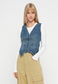 Τζιν γιλέκο με V-λαιμό, άνετο σχέδιο χωρίς μανίκια, με μπροστινά κουμπιά, δύο πλαϊνές τσέπες και κοντό τελείωμα. Φορεμένο πάνω από ένα λευκό μπλουζάκι με μακριά μανίκια.