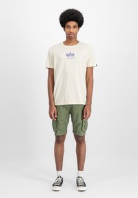 Beež puuvillane T-särk lilla logoga, kombineeritud roheliste cargo-lühikeste pükstega. Lühikesed püksid sisaldavad küljetaskuid ning neil on lõtv sobivus ja pikkus.