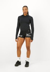 Melna sporta krekls ar garām piedurknēm un kapuci, apvienots ar melniem šortiem ar baltu "Nike Pro" tekstu. Modelis valkā melnus sporta apavus un baltas zeķes.
