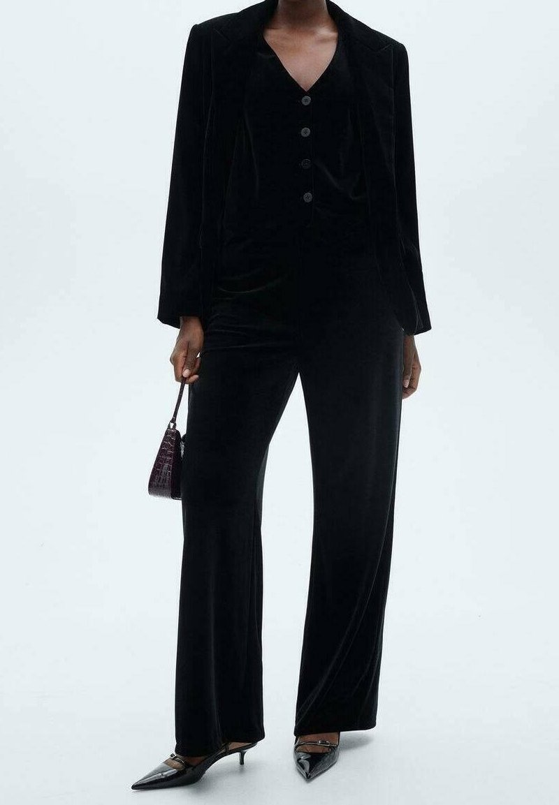 Femme portant un blazer en velours noir boutonné et un pantalon large, tenant un petit sac à main sombre et portant des talons noirs pointus.
