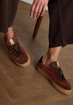 Sneakers gemaakt van gemengde materialen met bordeauxrood en bruin suede, zwarte veters en een dikke beige zool; luipaardprintaccenten aan de zijkanten.