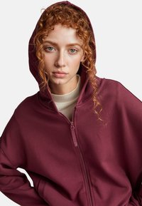 Haut à capuche zippé bordeaux avec cordon de serrage, présentant une texture lisse et une coupe décontractée. L'intérieur montre un col roulé crème superposé en dessous.