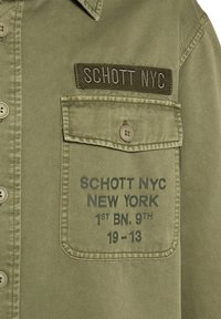 Poche de veste vert olive avec rabat à bouton, écusson brodé "SCHOTT NYC" et texte imprimé "SCHOTT NYC NEW YORK 1st BN. 9th 19-13."