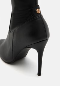 Bota de tacón alto de cuero negro con punta afilada, que presenta un corte lateral y un botón de acento dorado cerca del tobillo. Textura suave y diseño elegante.