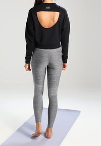 Czarny sweatshirt Nike z otwartym tyłem, w połączeniu z dopasowanymi, heatherowymi legginsami w odcieniu szarym. Modelka stoi na jasnej macie do jogi.