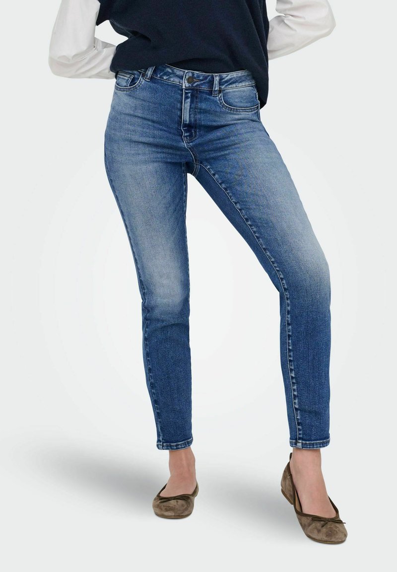 Femme portant un jean skinny bleu délavé, des ballerines beiges, un pull navy et une chemise blanche à manches longues, se tenant les jambes croisées.