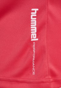 Rødt stof i nærbillede med "hummel" logoet og ordet "PERFORMANCE" i hvid, der fremhæver en glat tekstur og et rent design.