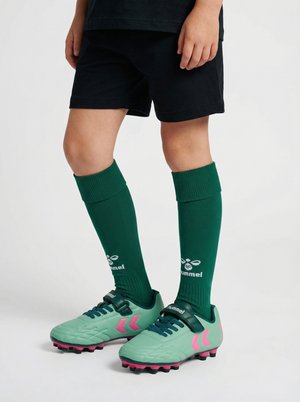 Dziecko noszące zielone korki piłkarskie z różowymi detalami, zielone sportowe getry do kolan i czarne spodenki na jednolitym jasnym tle.