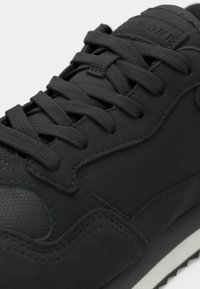 Musta lenkkari, jossa on sileä päällinen, teksturoidut yksityiskohdat ja litteät nauhat. Sisältää kumipohjan ja huomaamatonta brändäystä kielen kohdalla.