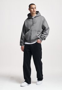 Tânăr cu păr scurt purtând un hanorac gri cu fermoar, blugi negri, adidași albi și un tricou alb, stând în fața unui fundal simplu.