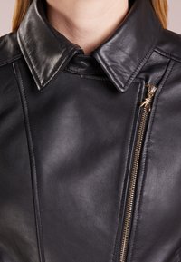 Mustat nahkatakki, jossa on teräväkauluksinen, vinossa olan vetoketju kullansävyisillä yksityiskohdilla ja sileä pinta. Ompeleet ovat näkyvissä.