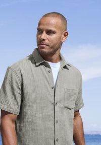 Camicia a maniche corte di colore grigio chiaro in tessuto texturizzato, con colletto, bottoni e taschino sul lato sinistro del petto, indossata sopra una camicia bianca.