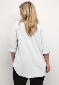 Witte, langharige shirt met opgerolde manchetten, zachte textuur, gebogen zoom en achterhalsdetail, gecombineerd met zwarte broeken.