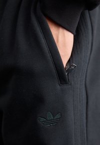 Sort athletic zip-up jakke med struktureret stof, sidelomme og præget logo. Glat overflade med kontrasterende syninger.