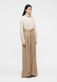 Γυναίκα με μακριά σκούρα μαλλιά, φορώντας μπεζ ζιβάγκο πουλόβερ και ριγέ μπεζ παντελόνια ψηλόμεσα με φαρδιά μπατζάκια, και καφέ παπούτσια, στέκεται πλάγια.