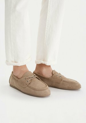 Füße in beige Wildleder-Halbschuhen mit Schnürsenkeln, kombiniert mit weißen hochgekrempelten Hosen auf weißem Hintergrund.