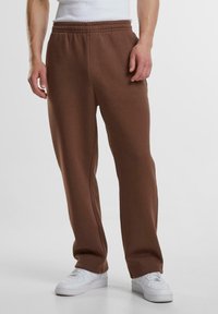 Brune sweatpants med elastisk talje, lige ben og sidelommer, parret med hvide sneakers på en ensfarvet baggrund.