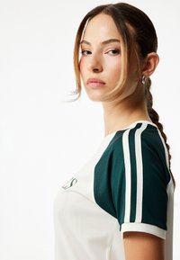 Urheilupaita, jossa on valkoinen pääosa ja vihreät hihat. Ominaisuuksiin kuuluu teksturoitu kangas ja valkoiset raidat hihoissa.