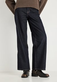 Σκούρο μπλε τζιν παντελόνι με φαρδιά γραμμή από denim ύφασμα, με αντιθετικές ραφές και ψηλή μέση, συνδυασμένο με μαύρα μποτάκια αστραγάλου.