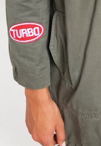 Brazo y mano de una persona que lleva una chaqueta verde oliva con un parche rojo y blanco que dice "TURBO" en la manga cerca de la muñeca.